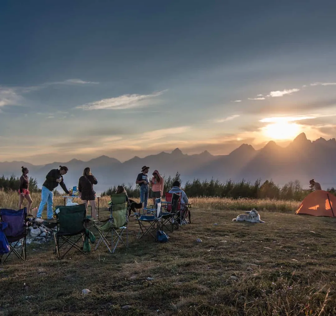 Campers gathered in a meadow as the sun sets behind the Tetons